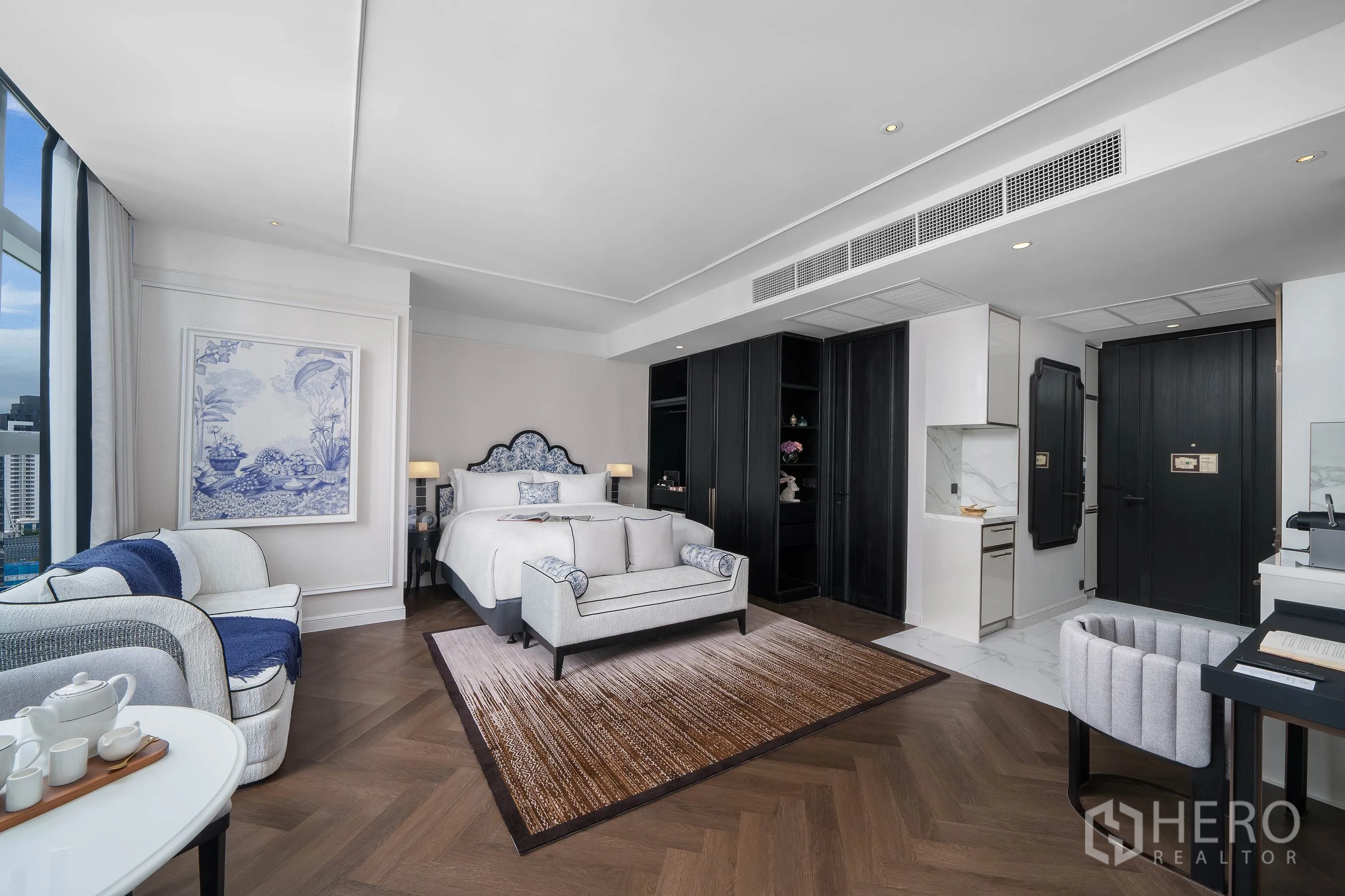 Condominium for rent in Khlong Toei, Bangkok - Spacious bedroom with sofa, rug, kitchenette, dark wood wardrobes, and city-facing windows.