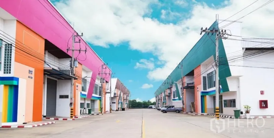 Warehouse for rent in Mueang Samut Sakhon, Samut Sakhon - Row of modern, colorful warehouse units with wide driveway, roller shutter doors, and overhead power lines in an industrial complex.