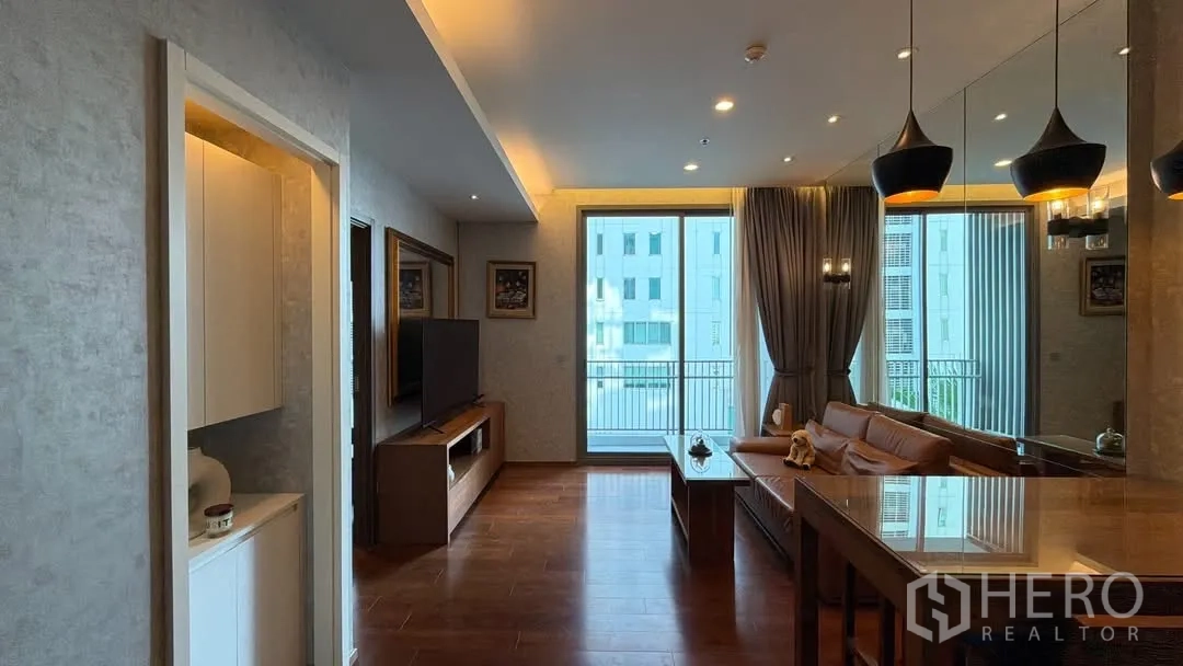 Condominium for rent in Watthana, Bangkok - Warm-toned living and dining area with leather sofa, pendant lights, wooden floors, TV console, and sliding glass doors opening to a balcony.