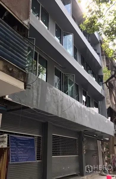 อาคารพาณิชย์ ให้เช่าในKhlong Toei, Bangkok - อาคารพาณิชย์รีโนเวทใหม่หลายชั้น ภายนอกสีเทา กระจกบานใหญ่ และประตูม้วนชั้นล่าง ใต้ร่มไม้บนย่านสุขุมวิท