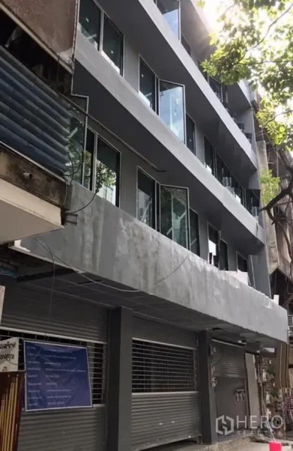 อาคารพาณิชย์ ให้เช่าในKhlong Toei, Bangkok - อาคารพาณิชย์รีโนเวทใหม่หลายชั้น ภายนอกสีเทา กระจกบานใหญ่ และประตูม้วนชั้นล่าง ใต้ร่มไม้บนย่านสุขุมวิท