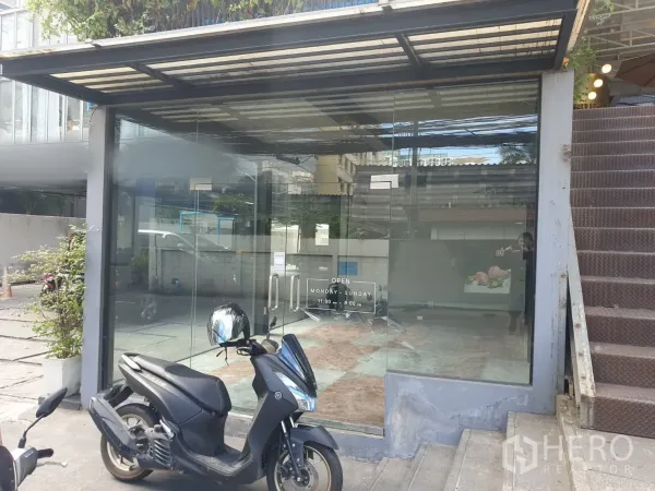零售空间 出租在Khlong Toei, Bangkok - 小巧店面，玻璃大门与金属雨棚相连，旁有楼梯，门前停放机车。