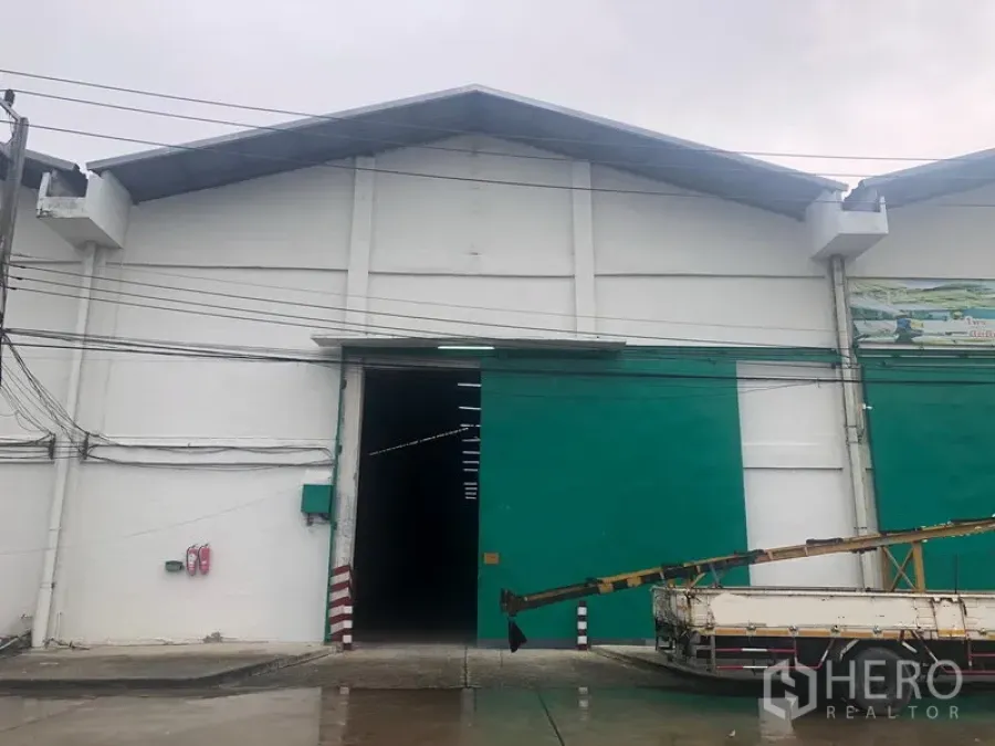 货栈 出租在Pathum Thani - 大型白色仓库外立面，绿色推拉门，卡车可进出。