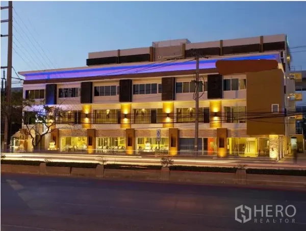 零售空间 出租在Khlong Toei, Bangkok - 傍晚的现代多层商业建筑，通透玻璃门面与暖色外立面灯光，临主干道醒目展示面，位于曼谷。