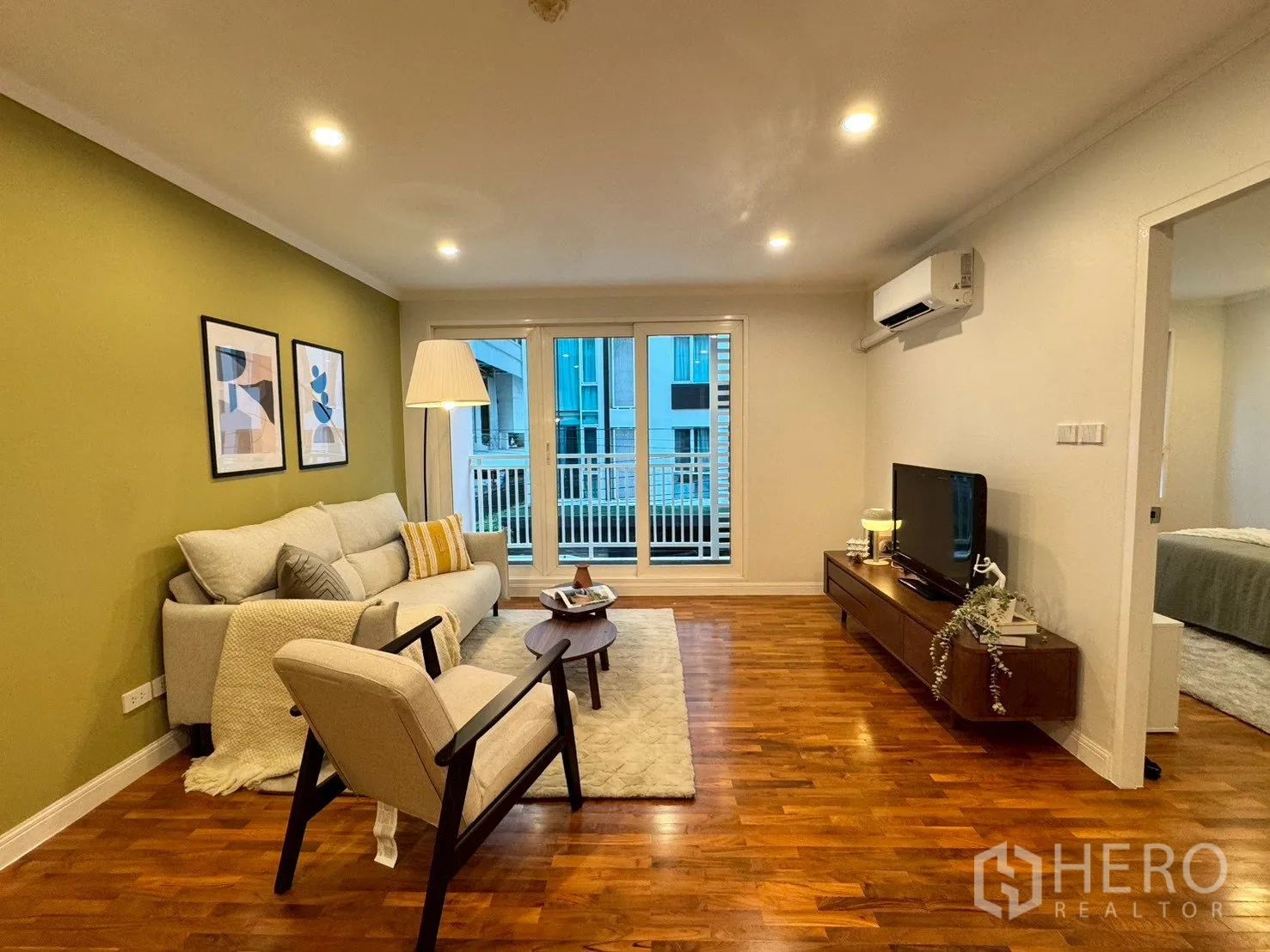 Condominium for sale in Watthana, Bangkok - Living room with sofa, armchair, TV console, wood floors, and sliding doors to a balcony with the bedroom doorway visible.