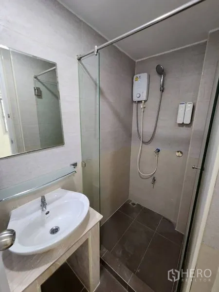 Home Office for rent in Khlong Toei, Bangkok - Compact bathroom with glass-enclosed shower, electric water heater, vanity sink, and gray wall tiles.