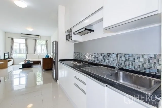 Townhouse for rent in Watthana, Bangkok - Sleek galley kitchen with white cabinetry, mosaic backsplash, double sink and view to living area.