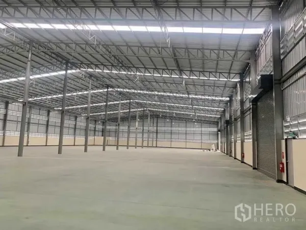 Warehouse for rent in Ban Bueng, Chon Buri - Spacious warehouse interior with high steel truss roof, smooth concrete floor, roller shutter doors, and fire safety fixtures.