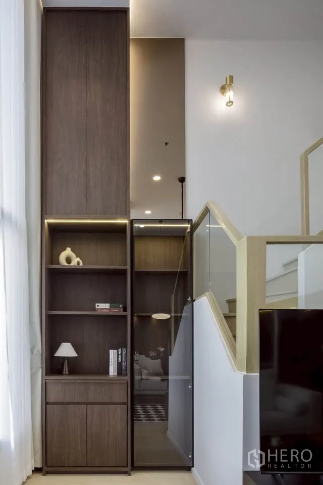 Condominium for rent in Khlong Toei, Bangkok - Staircase with glass railing beside tall built-in shelves and display lighting.