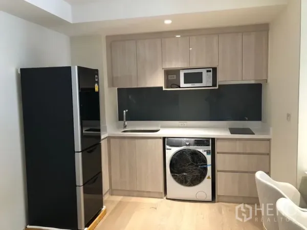 Condominium for rent in Watthana, Bangkok - Modern kitchenette with light wood cabinets, large black fridge, microwave and front-load washer.