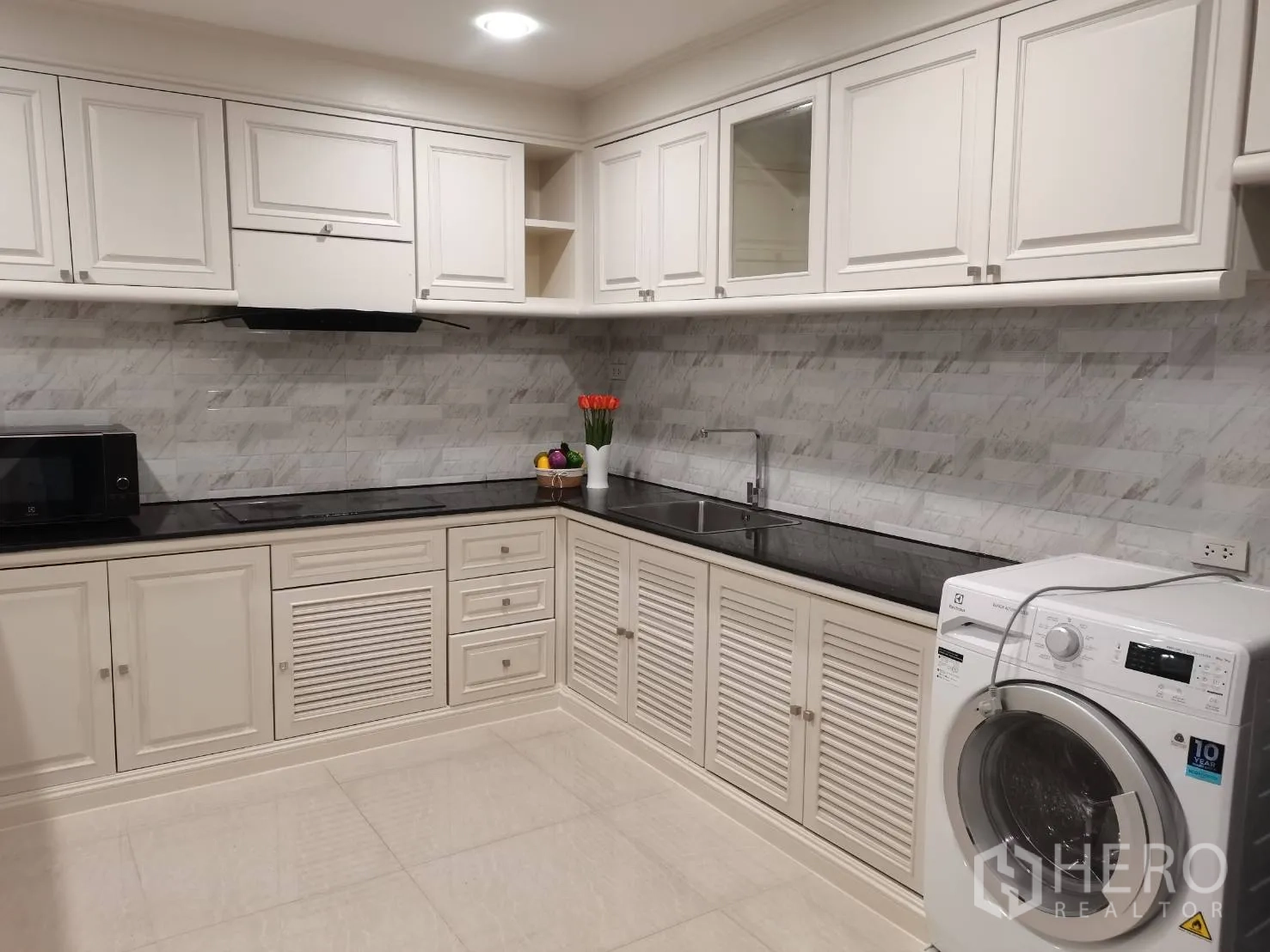 Condominium for rent in Watthana, Bangkok - White L-shaped kitchen with cooktop, hood, sink, microwave, and front-load washing machine against a tiled backsplash.