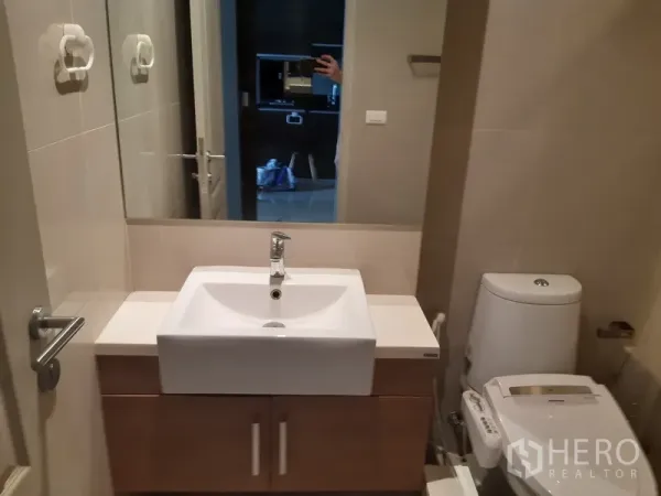 Condominium for rent in Bangkok - Compact bathroom with large mirror, white basin on wood vanity and modern toilet.
