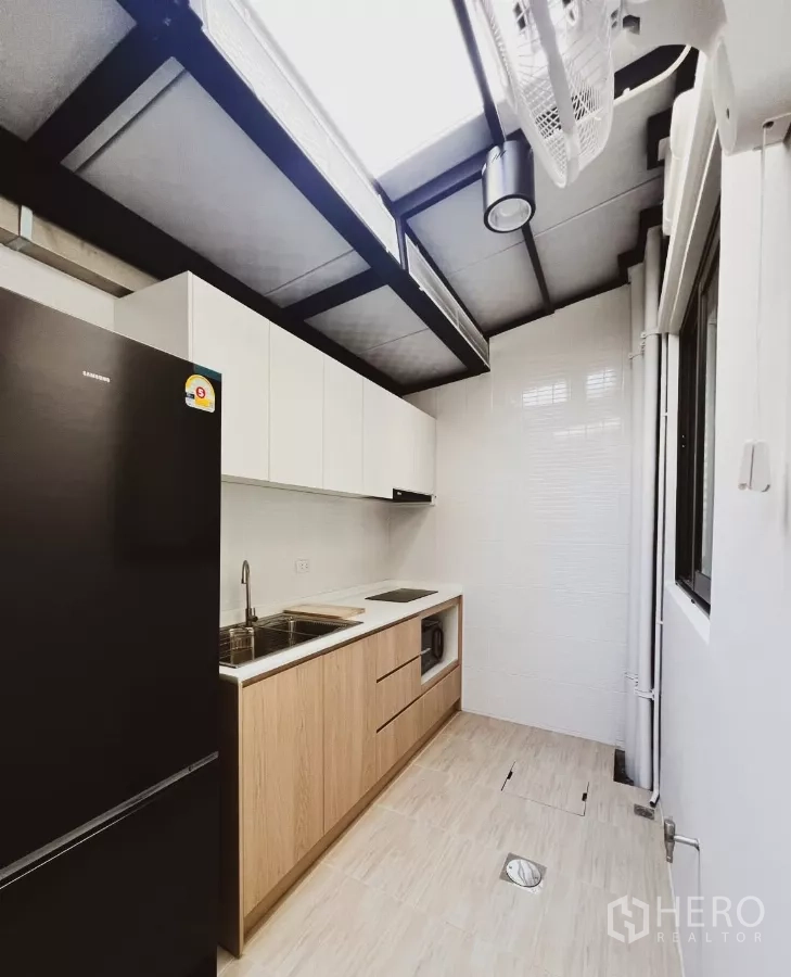 Townhouse for rent in Bang Phli, Samut Prakan - Extended rear kitchen with built-in cabinets, sink, electric cooktop, microwave niche and large fridge beneath a skylight.