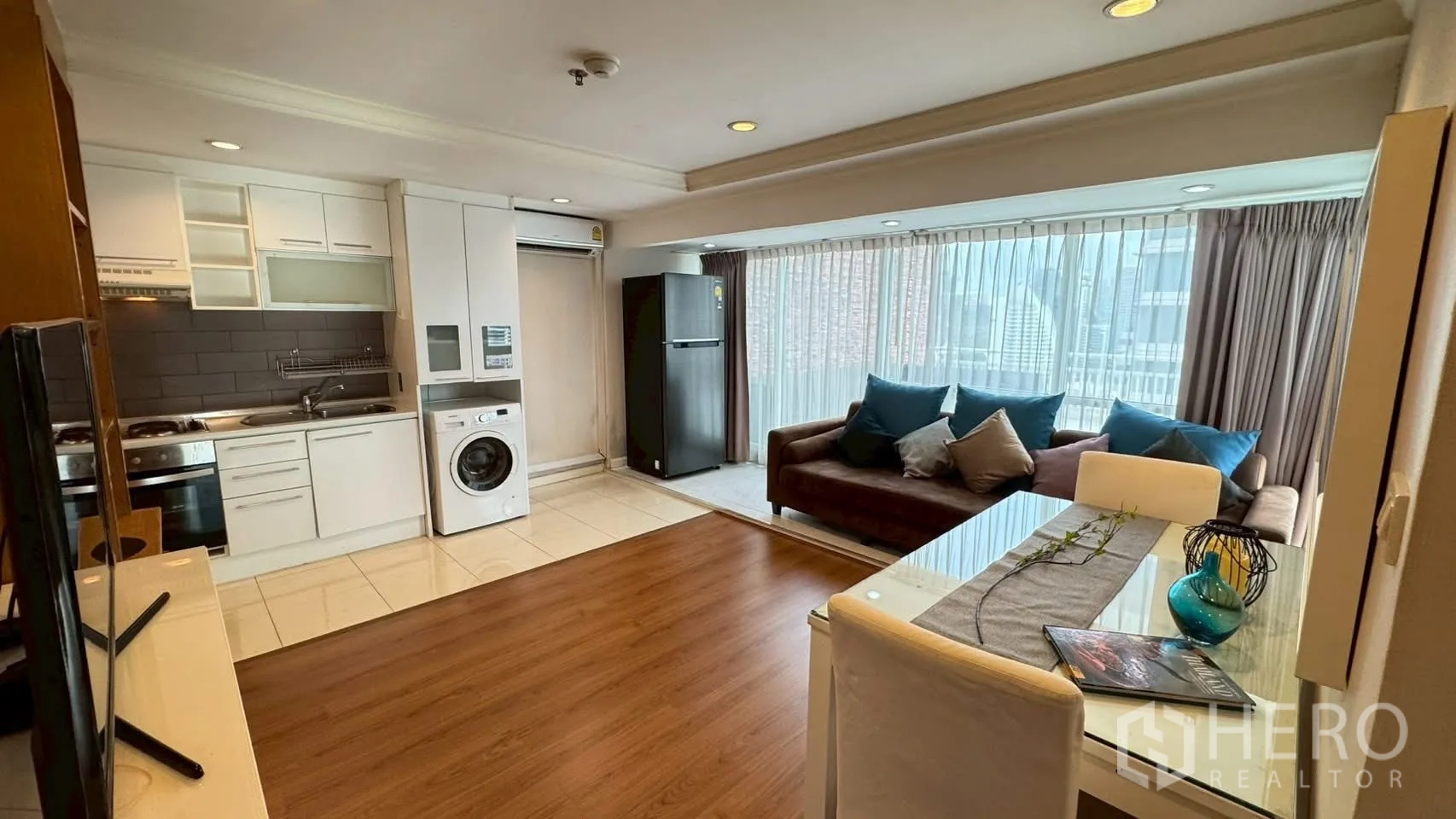 Condominium for rent or sale in Watthana, Bangkok - Open-plan living area with kitchenette, fridge, washing machine and sofa by large curtained windows.