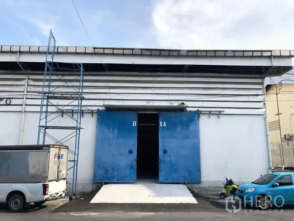 คลังสินค้า ให้เช่าในBang Phli, Samut Prakan - ด้านหน้าโกดัง ผนังสีขาว ประตูสไลด์เหล็กสีน้ำเงิน พร้อมทางลาดคอนกรีต และพื้นที่จอดรถด้านหน้า