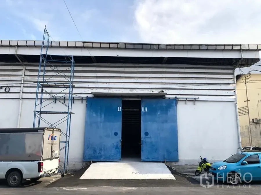 คลังสินค้า ให้เช่าในBang Phli, Samut Prakan - ด้านหน้าโกดัง ผนังสีขาว ประตูสไลด์เหล็กสีน้ำเงิน พร้อมทางลาดคอนกรีต และพื้นที่จอดรถด้านหน้า