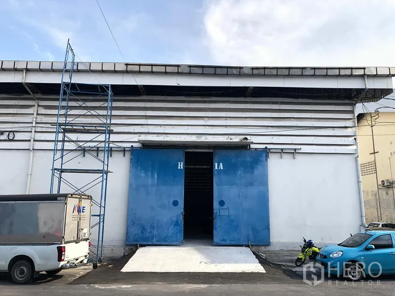 คลังสินค้า ให้เช่าในBang Phli, Samut Prakan - ด้านหน้าโกดัง ผนังสีขาว ประตูสไลด์เหล็กสีน้ำเงิน พร้อมทางลาดคอนกรีต และพื้นที่จอดรถด้านหน้า
