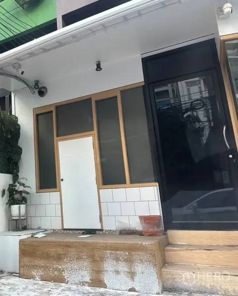 带办公室的家 出租在Watthana, Bangkok - 一层入口近景，黑色大门、磨砂窗、木质边框与监控摄像头。