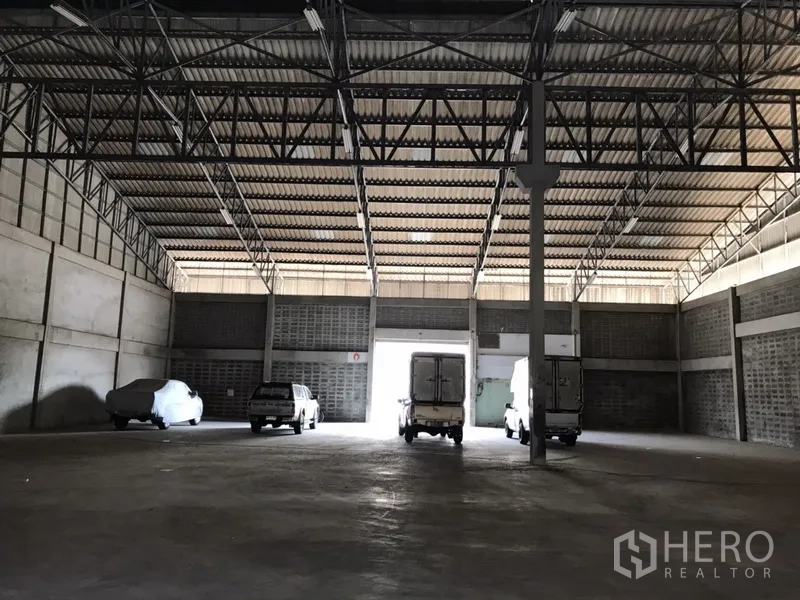 Warehouse for rent in Mueang Samut Prakan, Samut Prakan - Spacious warehouse interior with high steel-truss roof, concrete floor, and delivery trucks at the loading bay.