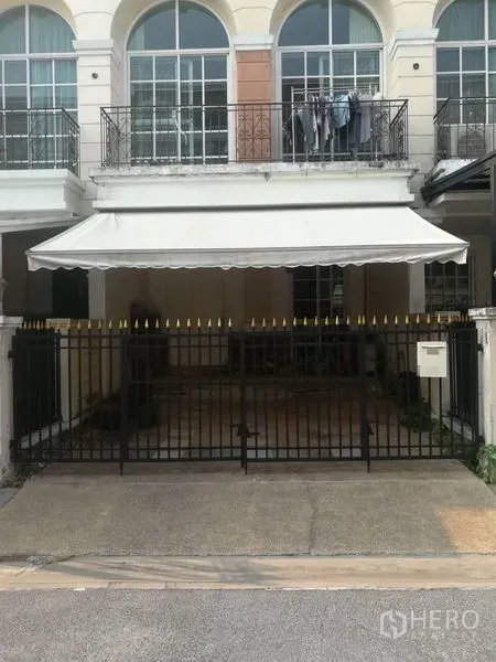 联排别墅 出售在Bang Kapi, Bangkok - 房屋正面，黑色金属栅栏与遮阳棚车库，楼上拱形落地窗。