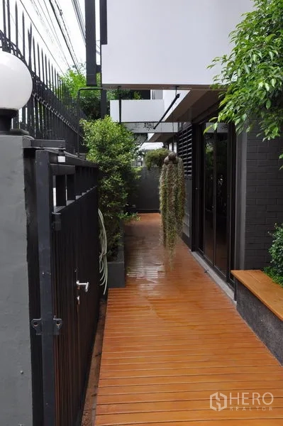 บ้าน ให้เช่าในWatthana, Bangkok - ทางเดินข้างบ้านพื้นไม้ ระเบียงยาว มีไม้ประดับห้อยและประตูรั้วสีดำปลอดภัย