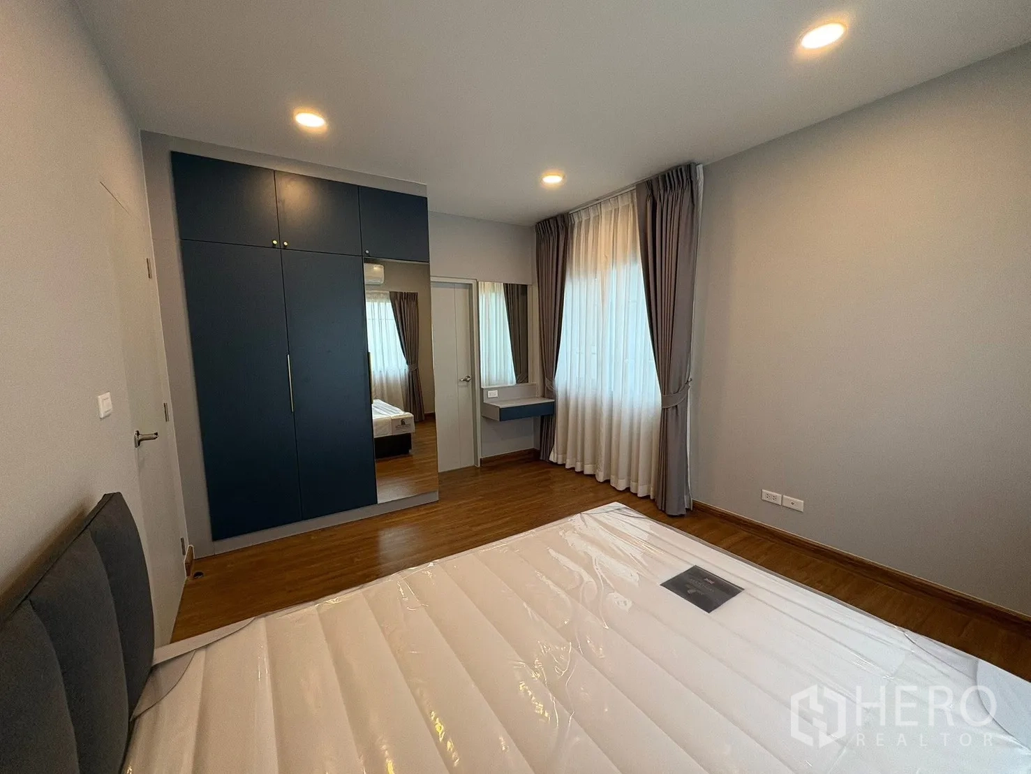 Detached House for rent in Bang Phli, Samut Prakan - Bright bedroom with a bed base, blue built-in wardrobe with mirrored door, vanity and curtains.