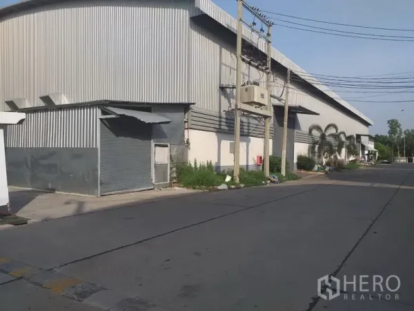 工厂 出租在Phra Nakhon Si Ayutthaya - 金属板外墙的仓库侧视图，设卷闸门，街侧有管线与电杆。