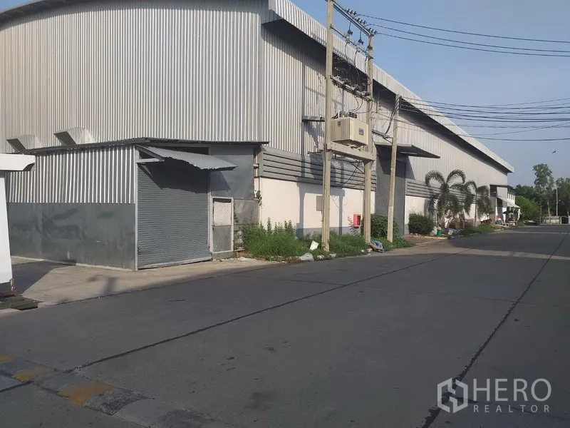 工厂 出租在Phra Nakhon Si Ayutthaya - 金属板外墙的仓库侧视图，设卷闸门，街侧有管线与电杆。