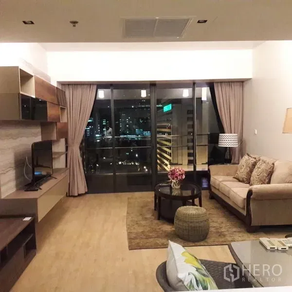 Condominium for sale in Sathon, Bangkok - Living area at night with sliding doors to a balcony framing city lights and a built‑in TV wall.
