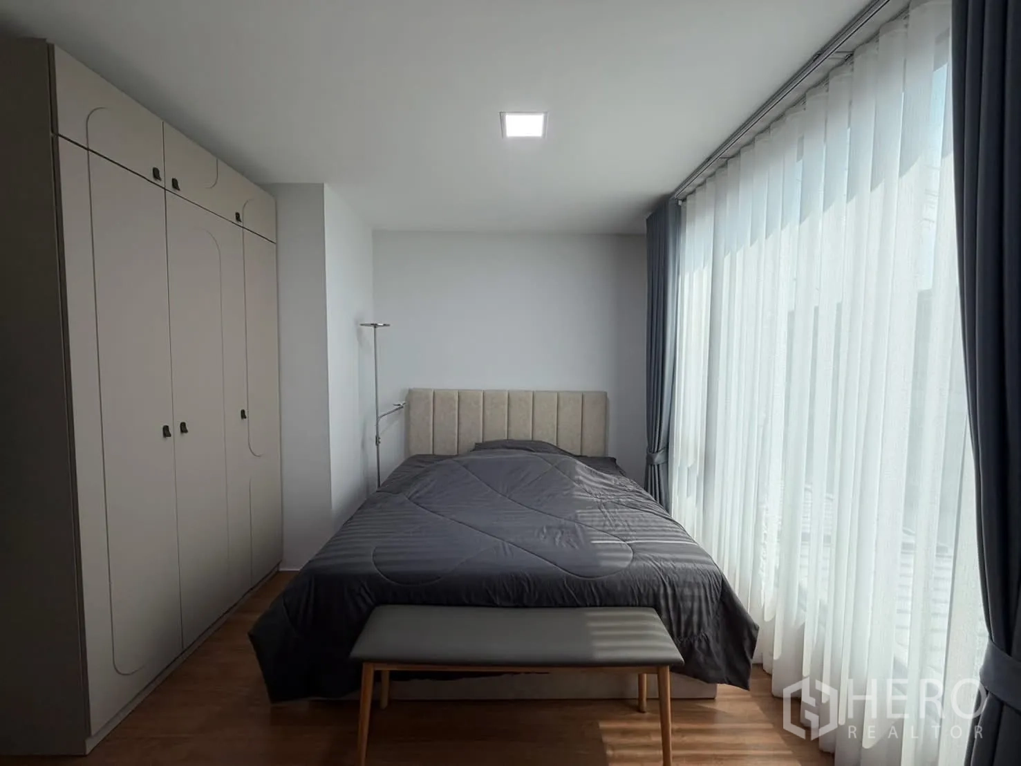 Townhouse for rent in Bang Phli, Samut Prakan - Primary bedroom with floor-to-ceiling windows, sheer curtains, upholstered headboard and bench, plus ample wardrobes.