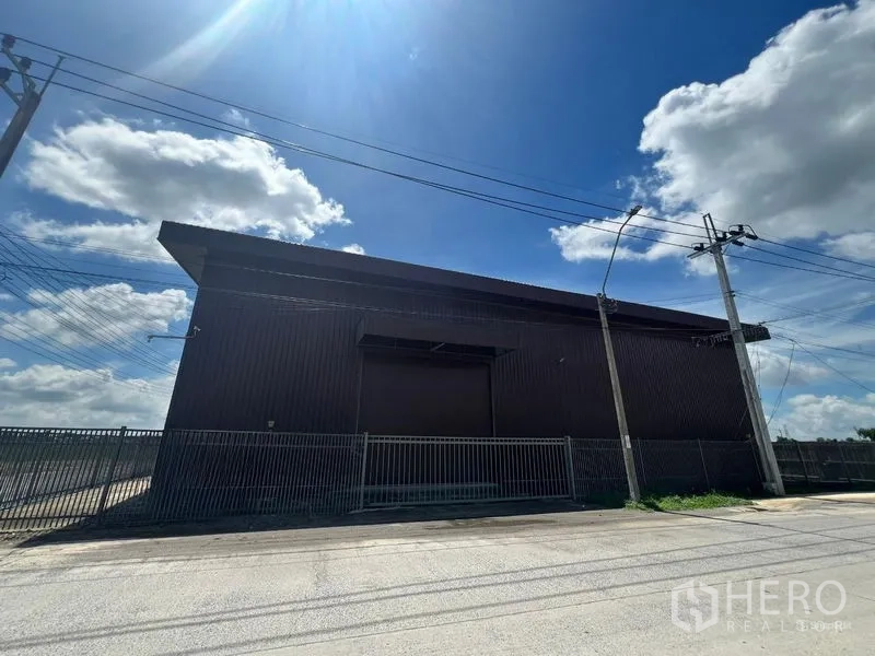 คลังสินค้า ให้เช่าในMin Buri, Bangkok - มุมมองด้านหน้าโกดังผนังเมทัลชีท พร้อมประตูม้วนและทางเข้าแบบมีรั้ว ใต้ท้องฟ้าสดใสในเขตมีนบุรี