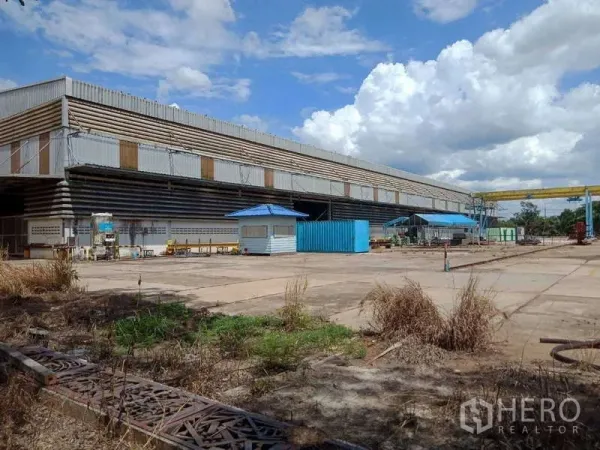 工厂 出售在Chachoengsao - 大型钢结构工厂，宽阔混凝土场地，蓝色门卫亭与门式起重机，天空多云。