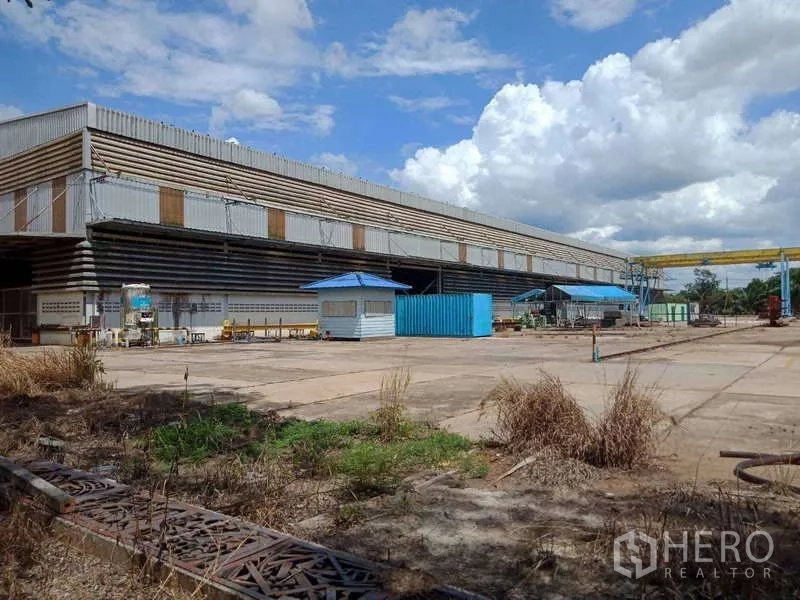 工厂 出售在Chachoengsao - 大型钢结构工厂，宽阔混凝土场地，蓝色门卫亭与门式起重机，天空多云。