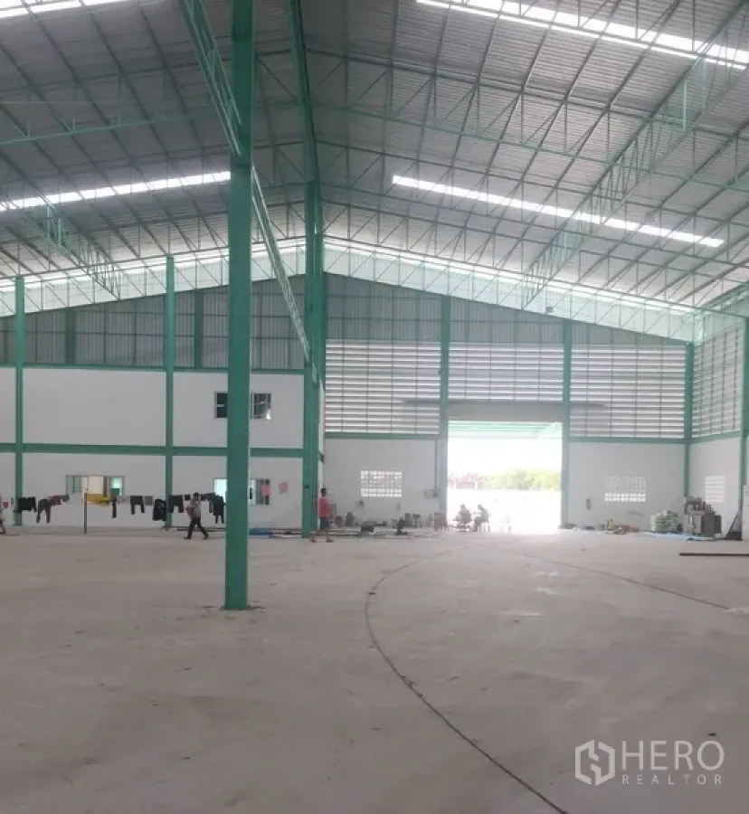 Warehouse for rent in Samut Prakan - Spacious high-clearance warehouse interior with green steel trusses, bright roof lights, and a large loading entrance.