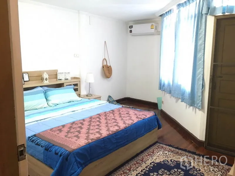 Townhouse for rent in Khlong Toei, Bangkok - Bedroom with blue curtains, wooden floor, double bed and bedside lamp.