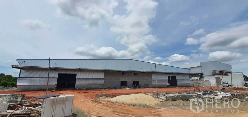 Factory for rent in Mueang Samut Sakhon, Samut Sakhon - Wide metal-clad factory exterior with loading bays and red earth yard under a cloudy sky in Samut Sakhon.