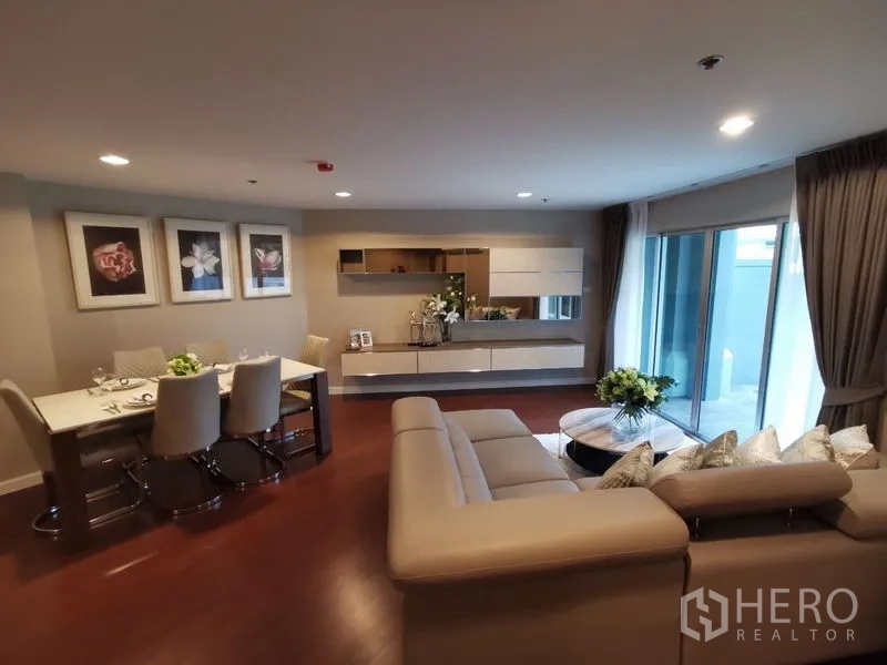 Condominium for rent or sale in Huai Khwang, Bangkok - Open-plan living and dining room with L-shaped sofa, sliding glass doors to balcony, and wall cabinets.