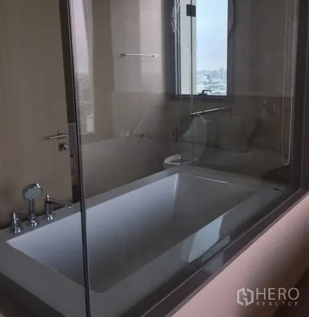 Condominium for rent in Watthana, Bangkok - Modern bathroom with glass‑enclosed bathtub and a window view of the city.