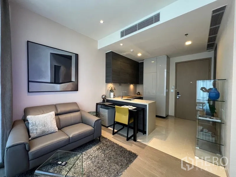 Condominium for rent in Watthana, Bangkok - Modern Bangkok condo living room with grey sofa, artwork, and a compact open kitchen with breakfast bar in the Asok–Sukhumvit area.