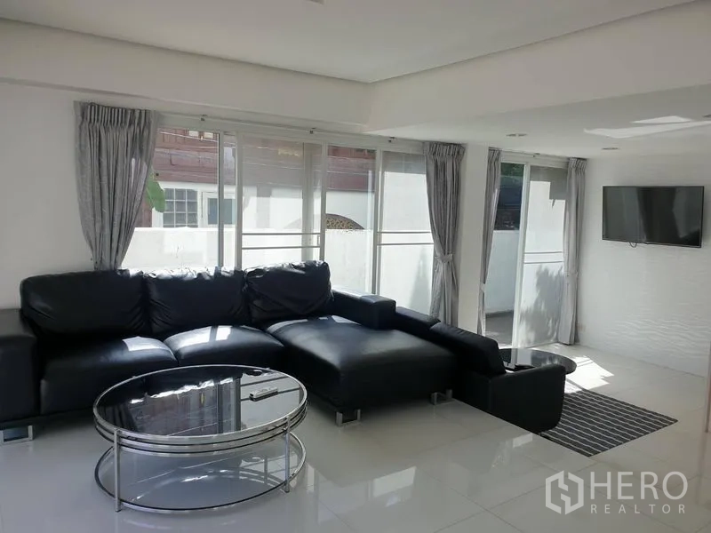 Detached House for rent in Khlong Toei, Bangkok - Bright living room with black leather sectional, glass coffee table and floor-to-ceiling windows.