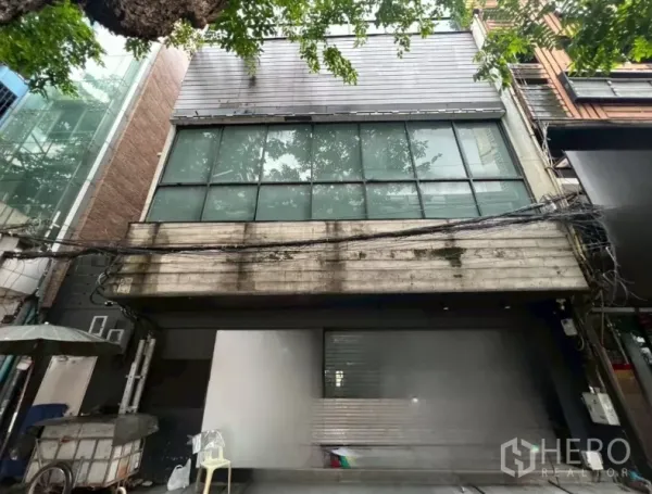 店屋 出租在Bang Rak, Bangkok - 临街商铺外立面，宽大玻璃窗与金属饰面，树荫掩映