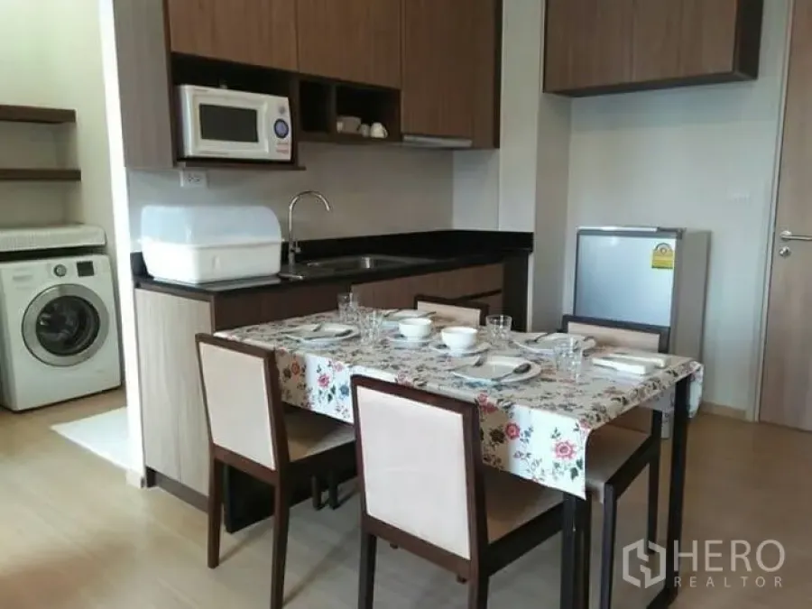 Condominium for rent in Huai Khwang, Bangkok - Compact kitchen and dining area with wood cabinetry, microwave, fridge and washing machine in a Bangkok condo.