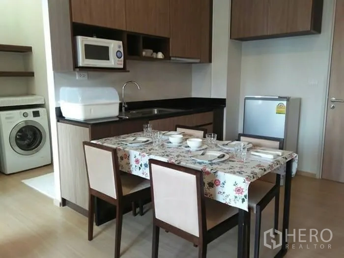 Condominium for rent in Huai Khwang, Bangkok - Compact kitchen and dining area with wood cabinetry, microwave, fridge and washing machine in a Bangkok condo.
