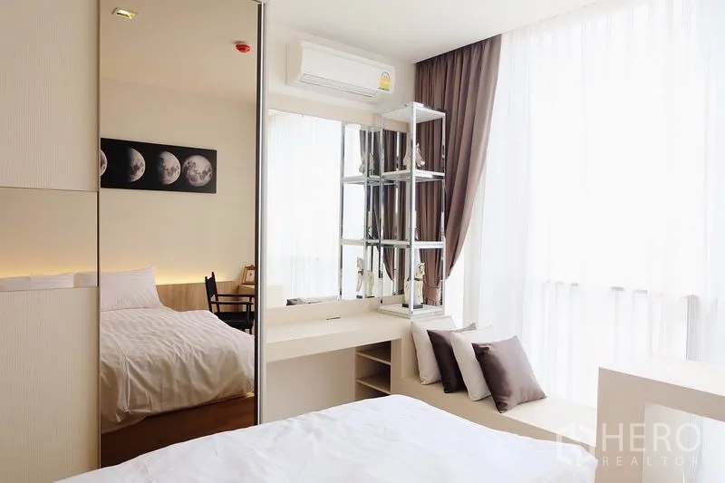 Condominium for sale in Watthana, Bangkok - Bedroom with mirrored wardrobe, vanity desk and bright curtains letting in natural light.