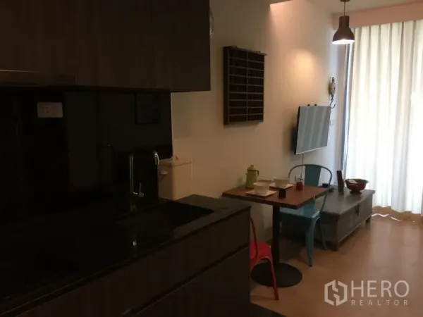 公寓 出租在Watthana, Bangkok - 紧凑厨房，深色橱柜与水槽，吊灯，小餐桌对着壁挂电视。