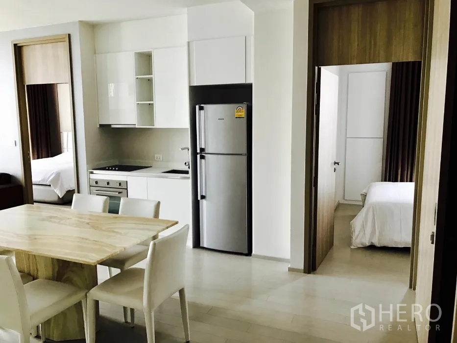 Condominium for rent or sale in Pathum Wan, Bangkok - Kitchen and dining zone with stainless refrigerator, showing doorways to two separate bedrooms.