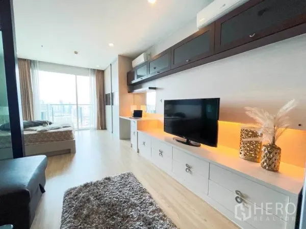 公寓 出租在Watthana, Bangkok - 开放式客厅，内置电视柜与书桌，推拉玻璃门通向阳台。