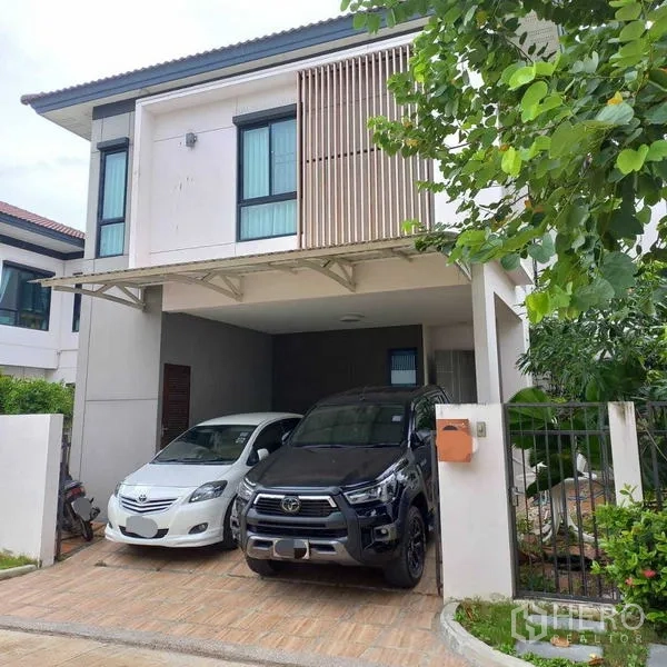 Detached House for rent in Bang Khun Thian, Bangkok - Front view of the home showing two vehicles under the covered carport, large upstairs windows and a small side garden.