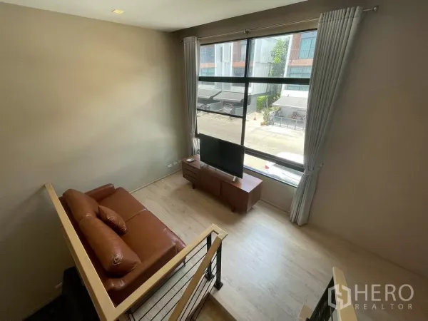 ทาวน์เฮาส์ ให้เช่าในSuan Luang, Bangkok - เลานจ์ชั้นลอยพร้อมโซฟาหนังสีน้ำตาล ชั้นวางทีวี และหน้าต่างบานกว้างมองเห็นซอยพักอาศัยเงียบสงบ.