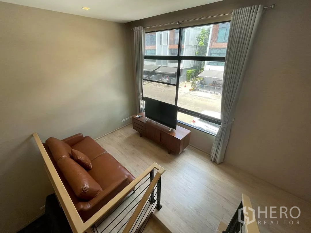 ทาวน์เฮาส์ ให้เช่าในSuan Luang, Bangkok - เลานจ์ชั้นลอยพร้อมโซฟาหนังสีน้ำตาล ชั้นวางทีวี และหน้าต่างบานกว้างมองเห็นซอยพักอาศัยเงียบสงบ.